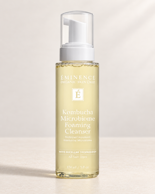 A bottle of Ŝminence Organic Skincare Kombucha Microbiome Foaming Cleanser, featuring a clear bottle filled with pale golden liquid and topped with a white pump dispenser, set against a warm beige background with soft shadows.
