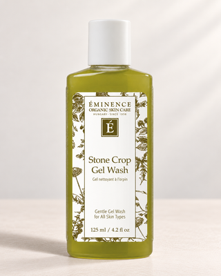 A bottle of Éminence Organic Skin Care Stone Crop Gel Wash, featuring a translucent olive-green liquid and a white cap, set against a clean, light beige background.