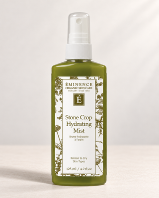 A bottle of Eminence Organic Skin Care Stone Crop Hydrating Mist in a translucent olive green bottle with a white spray top and a cream-colored label with botanical illustrations.