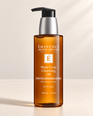 A bottle of Éminence Organic Skin Care Stone Crop Cleansing Oil with an amber brown body, a silver pump collar, and a black pump dispenser, set against a warm beige background with soft shadows.